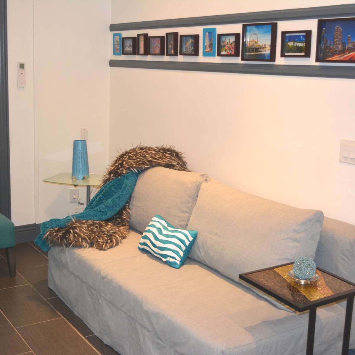 A living room with a gray sofa, a teal accent chair, a small glass side table, and a series of framed pictures mounted on the wall.
