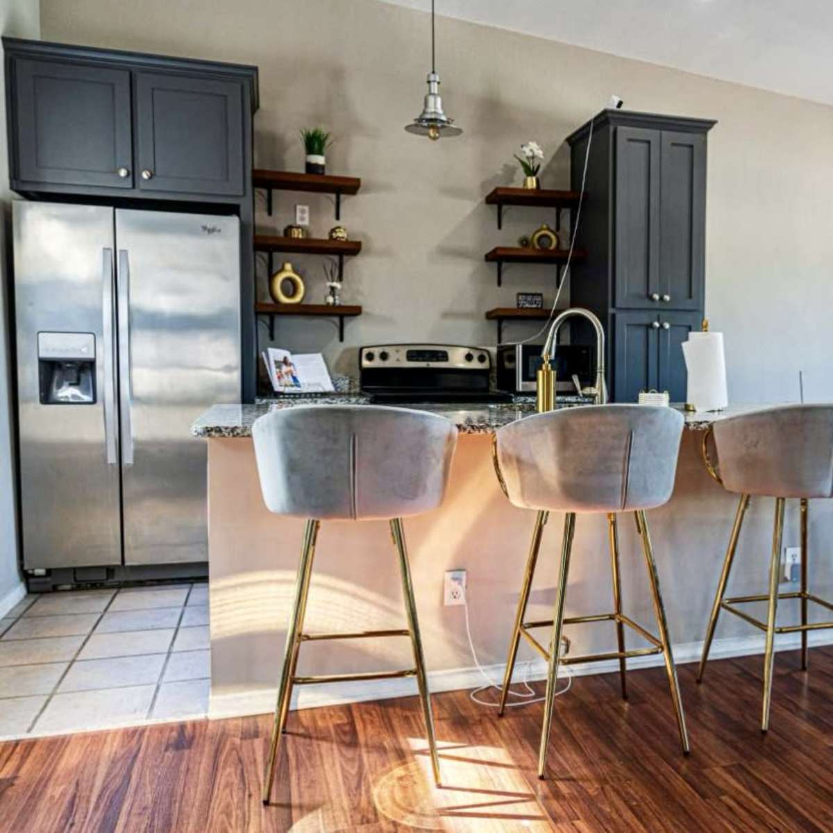 A modern kitchen with gray cabinetry, a stainless steel refrigerator, and four bar stools lined up at a granite-topped island.
