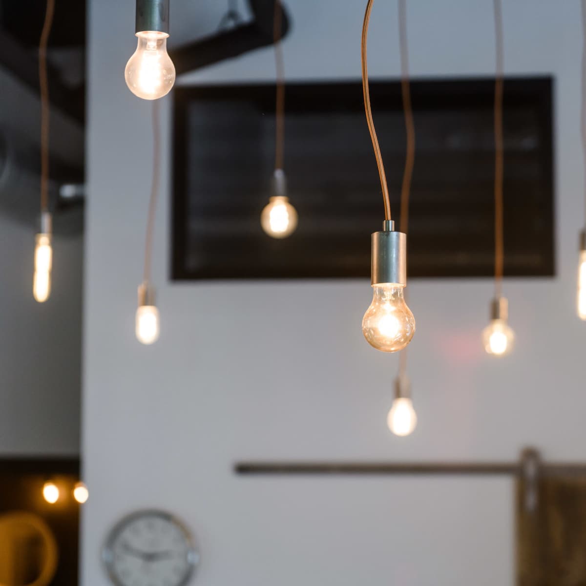 A group of hanging light bulbs with metal fixtures illuminates a room with a dark wall and a clock in the background.