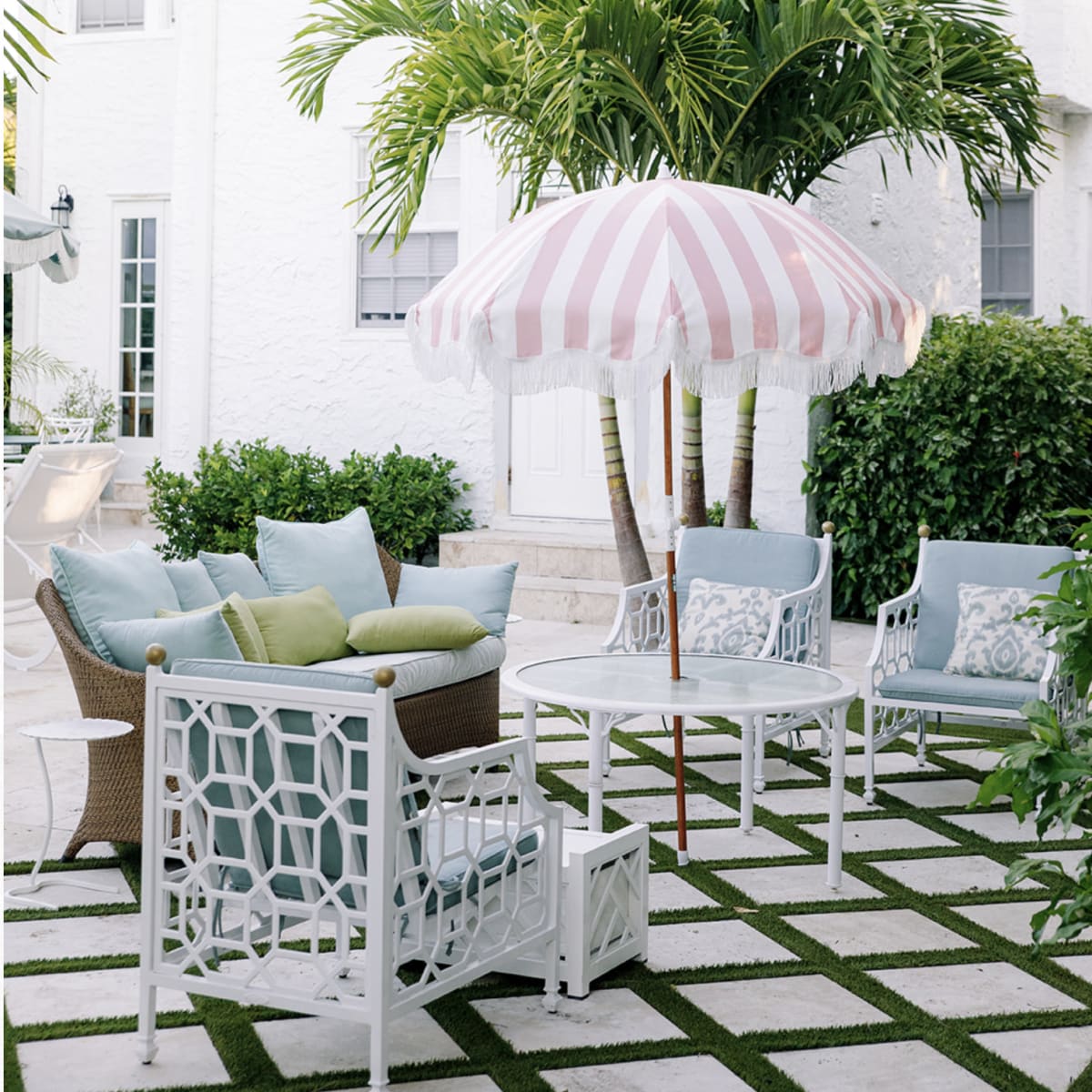 A cozy outdoor seating area features a round table with striped umbrella, surrounded by cushioned chairs and lush green foliage.