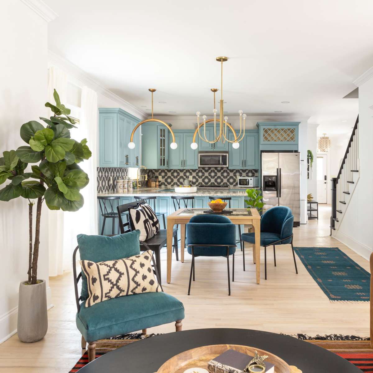 A modern open-concept living and dining area featuring blue cabinetry, a dining table with teal chairs, and a staircase in the background.