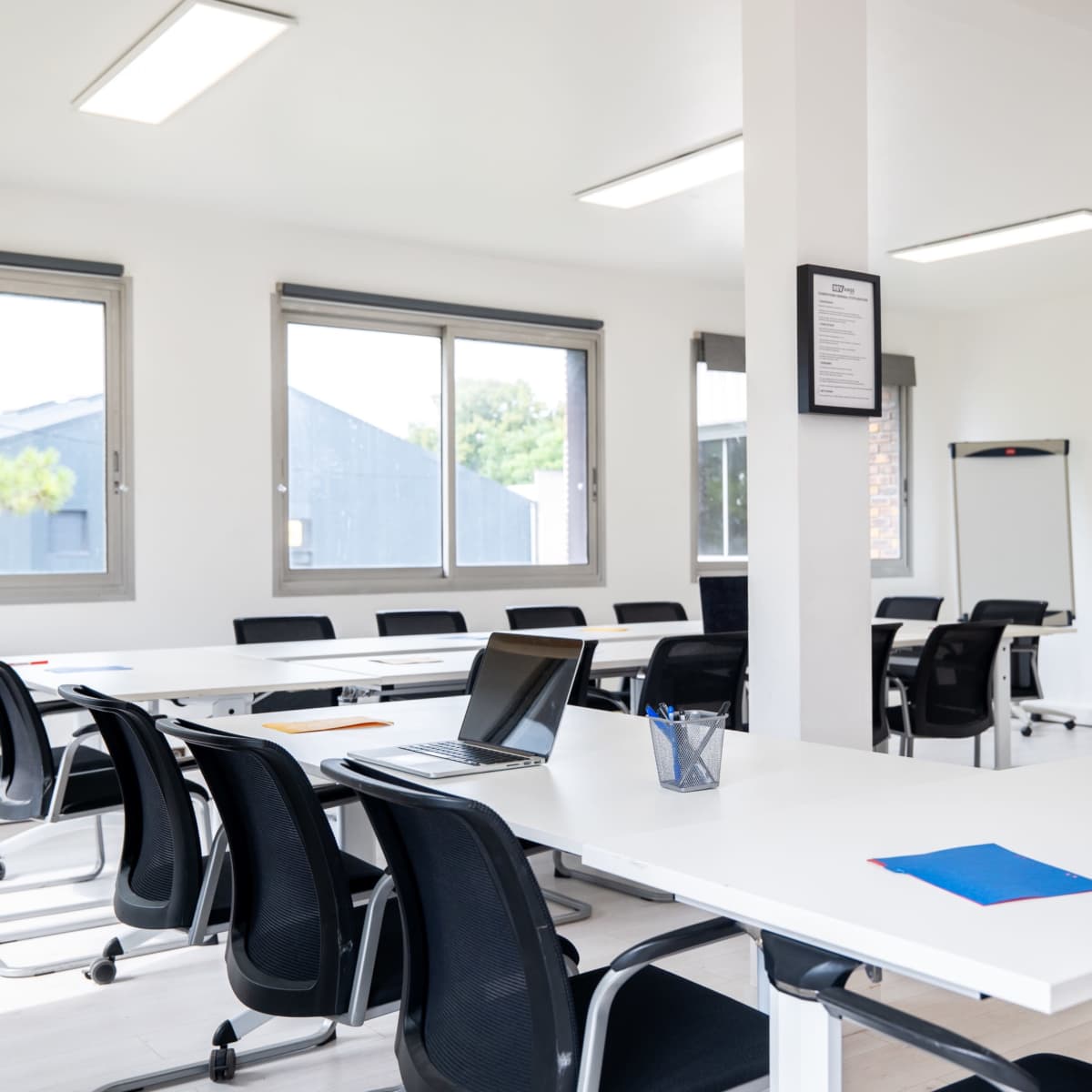 A modern meeting room features a long table with black chairs, a laptop, and colorful stationery, along with large windows allowing natural light.