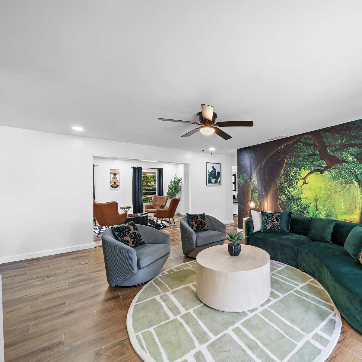 A modern living room featuring a curved green sofa, two armchairs, a round coffee table, and a mural of a forest on one wall, with a potted plant in the corner.