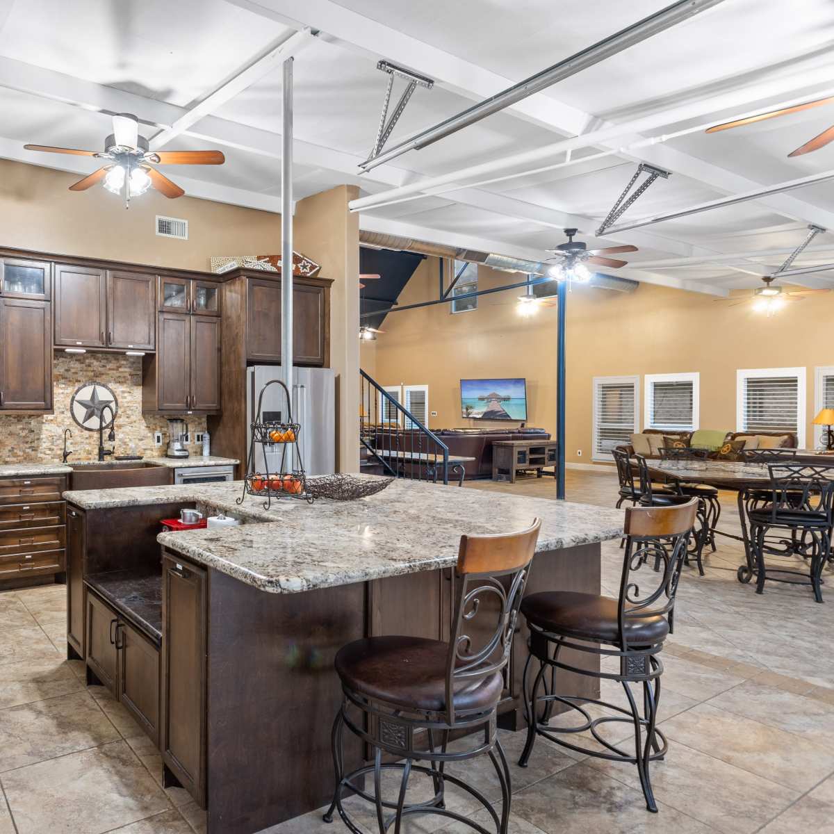 A spacious kitchen with dark wooden cabinets, granite countertops, and an island with bar stools, adjacent to a large open living area.