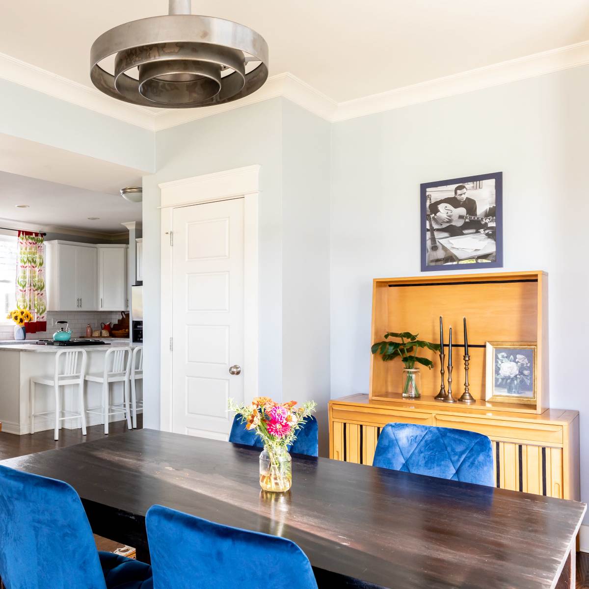 A dining area with a large dark table and blue chairs, leading into a kitchen with white cabinetry and a mirror on the wall.