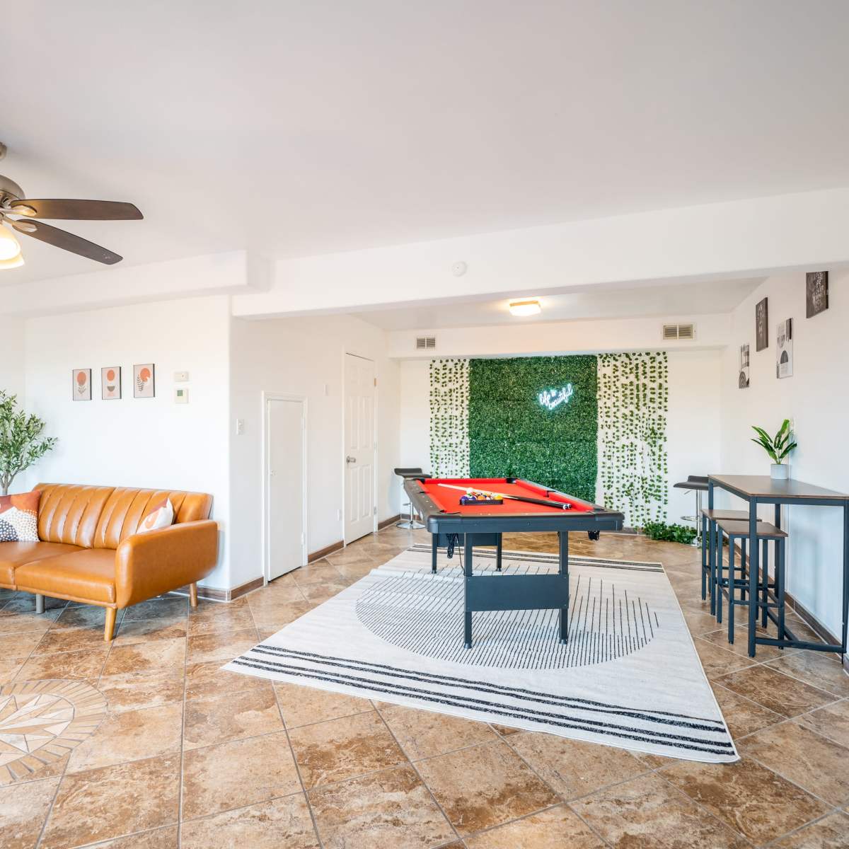 A well-lit living space with a pool table, a brown leather sofa, and a decorative wall covered in green foliage.
