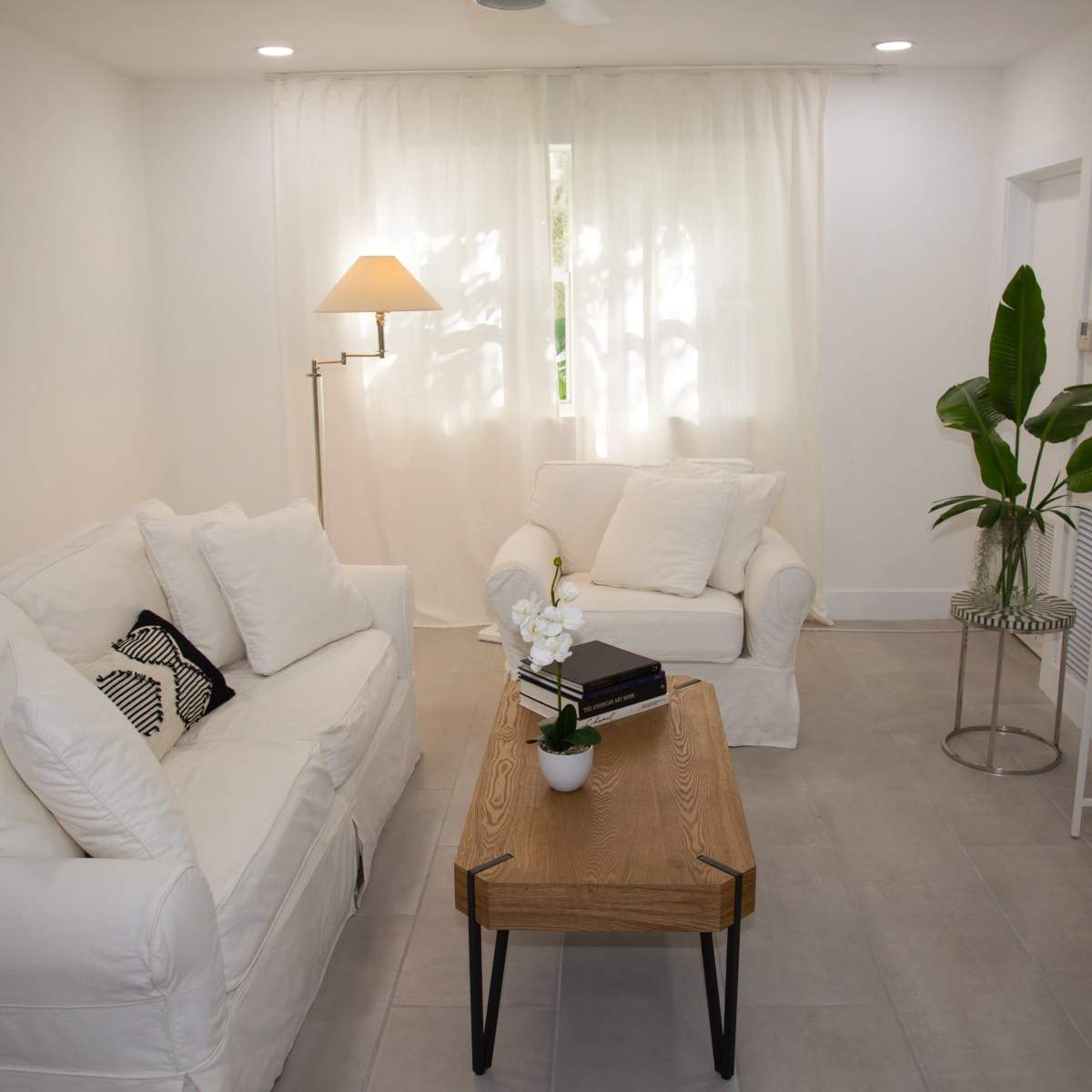 A bright living room features two white sofas, a wooden coffee table, a plant, and a television.