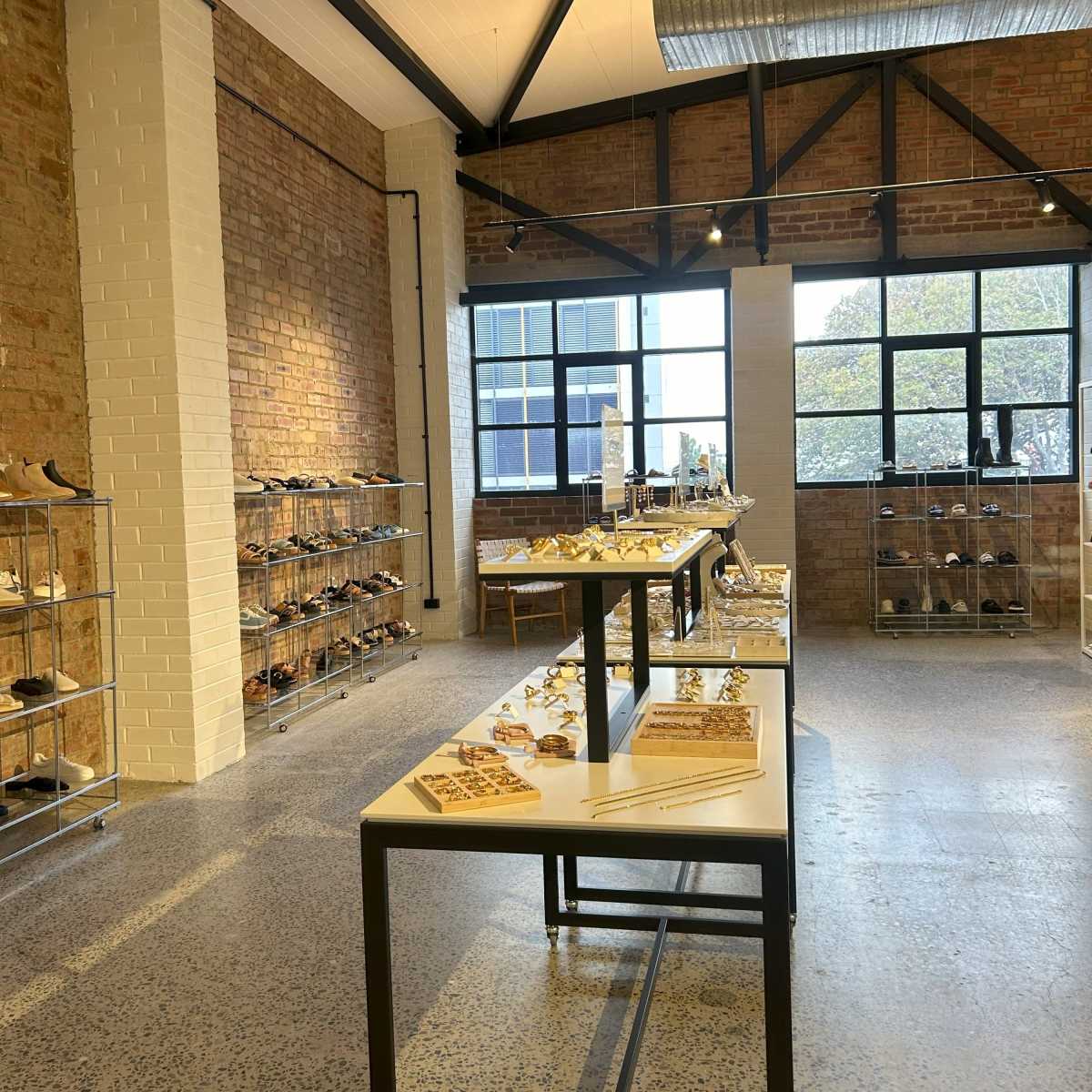 A shoe store interior with multiple display tables and shelves showcasing various footwear, set against a backdrop of brick walls and large windows.