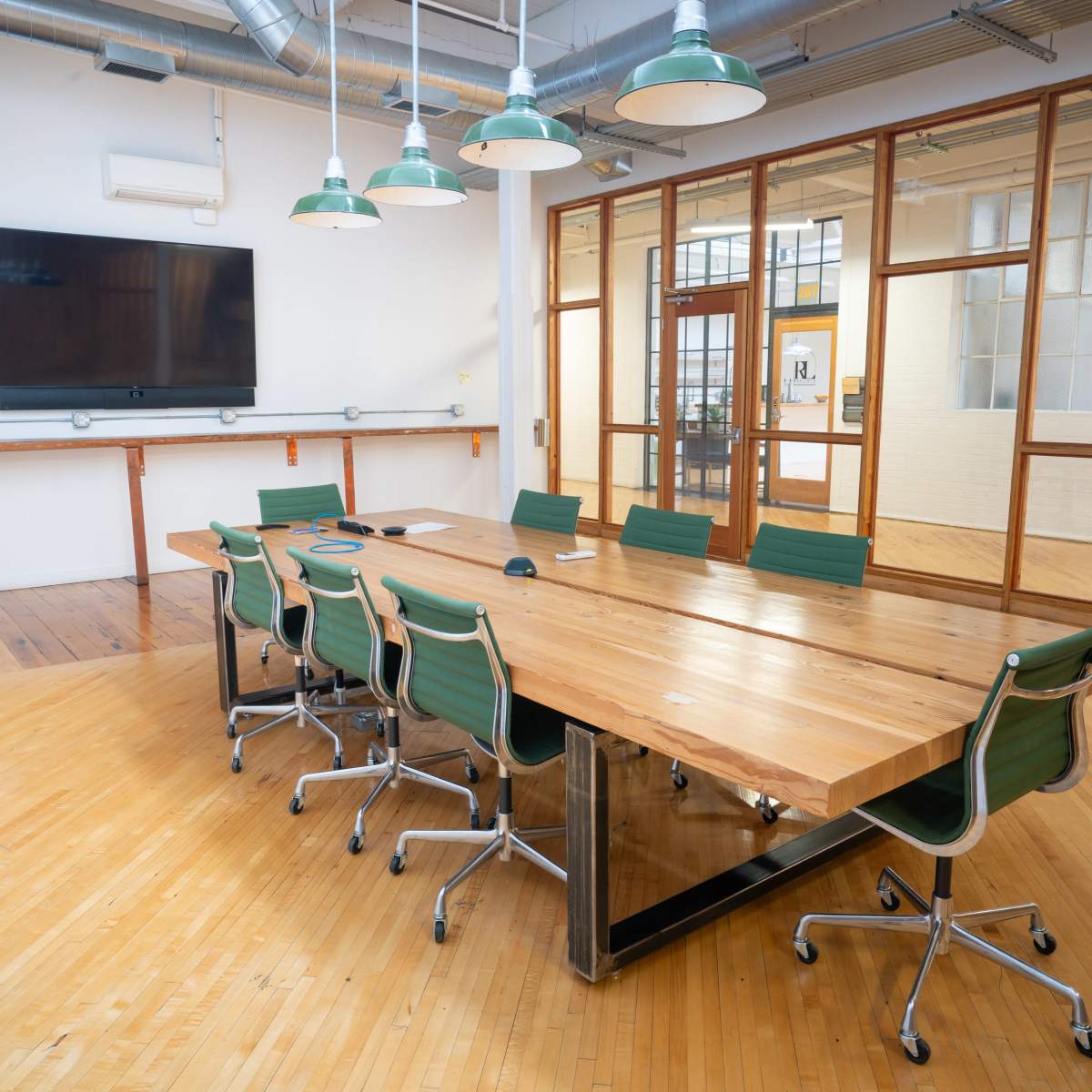 A large wooden conference table with green chairs is positioned in a bright room featuring a TV mounted on the wall and large glass doors.