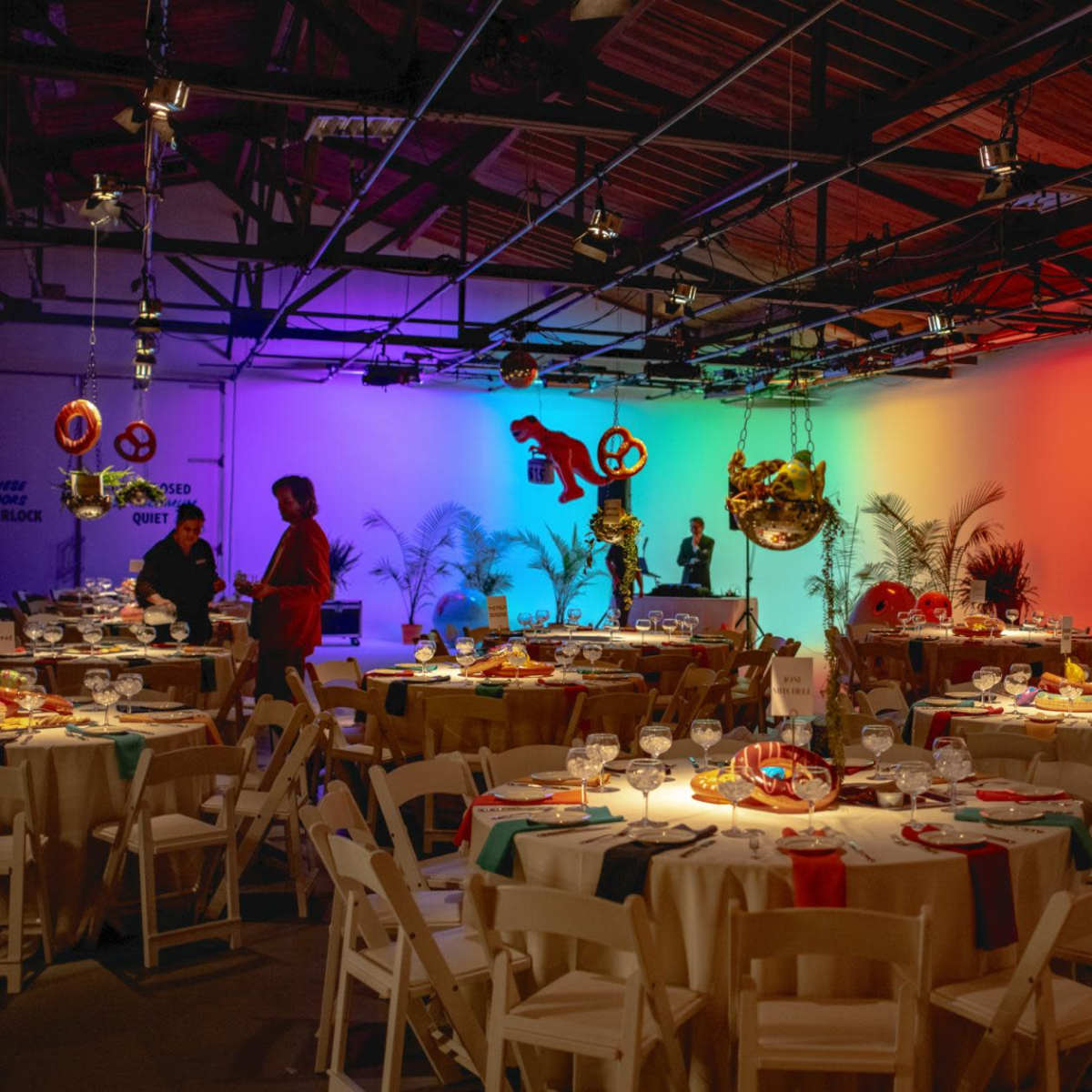 A vibrant event space with round tables set for a banquet, colorful lighting illuminating the background, and decorative elements hanging from the ceiling.