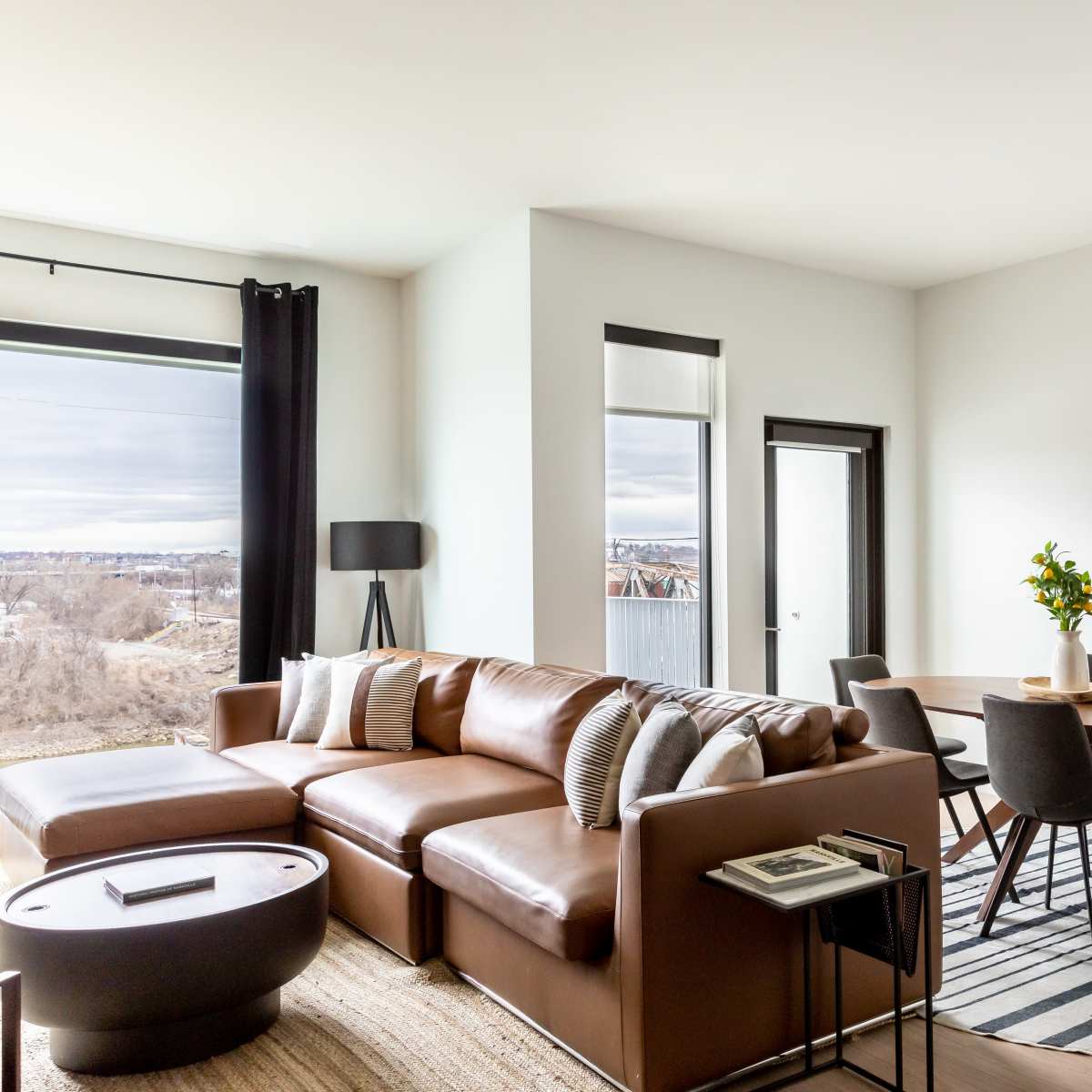 A modern living room and dining area with a brown sectional sofa, round dining table, and large windows overlooking a scenic view.