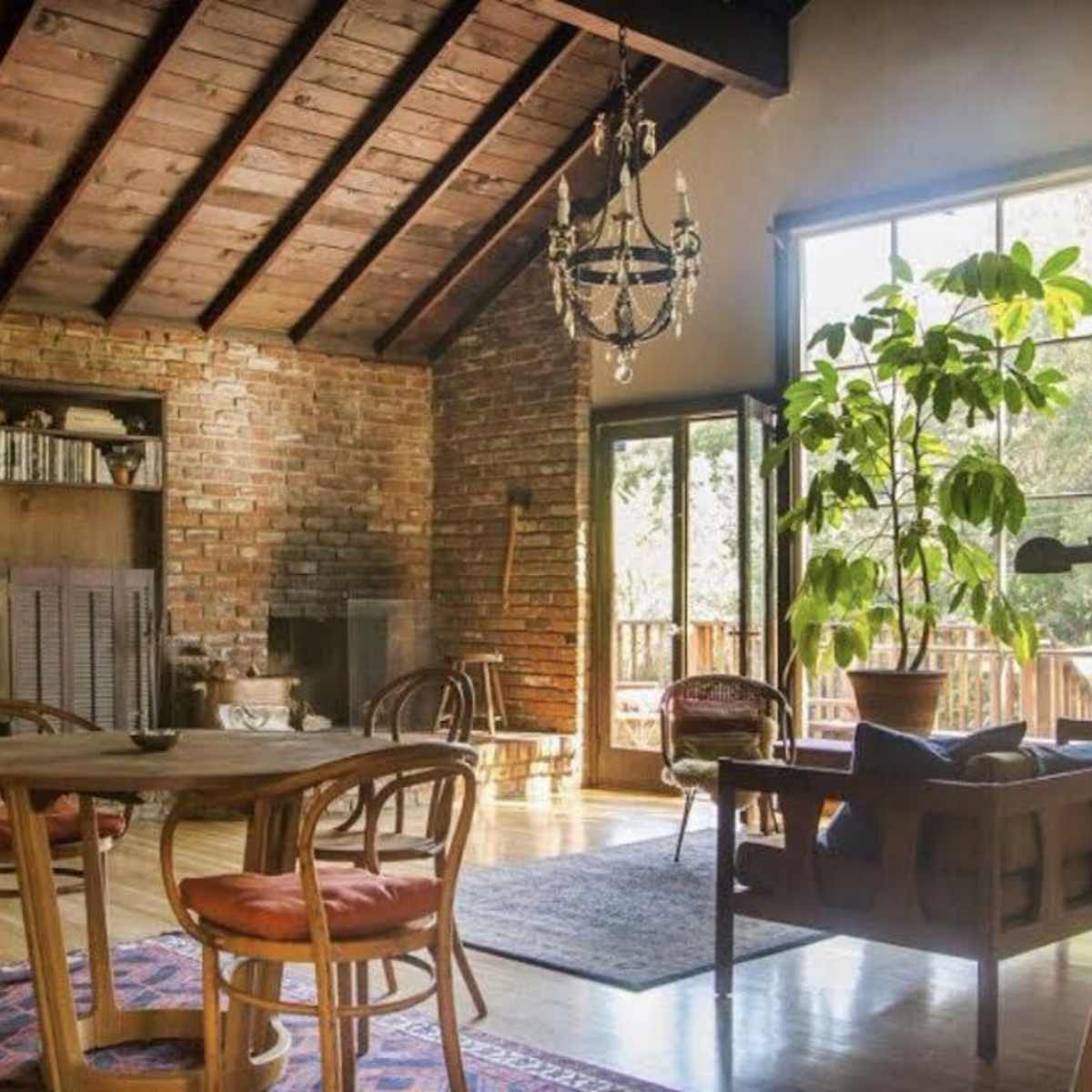 A cozy living room features a wooden ceiling, a brick wall, a dining table, and large windows with natural light streaming in.