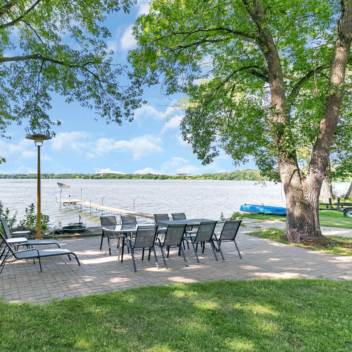 A patio area with outdoor seating and a lamp overlooks a lake, flanked by trees and a dock.