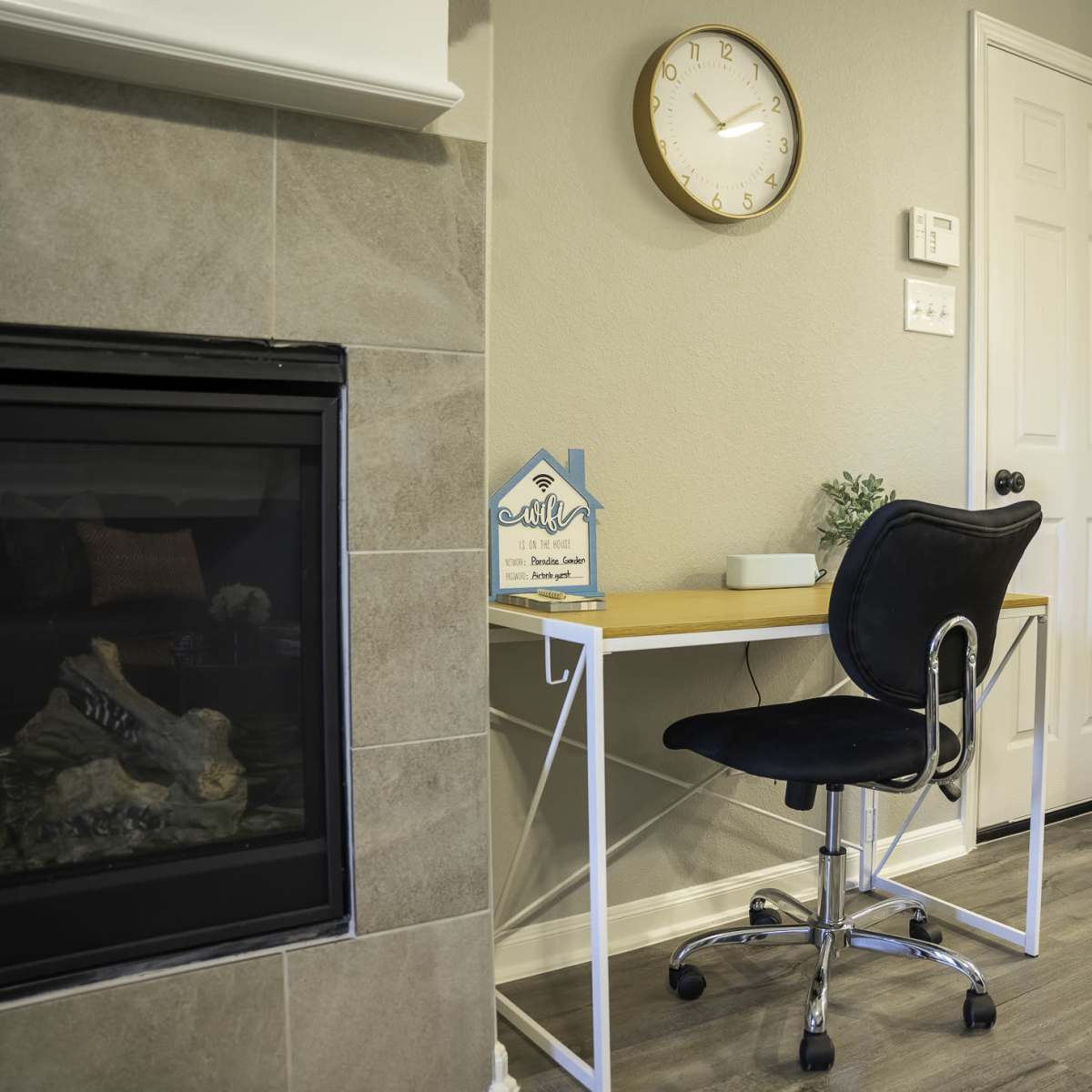A modern home office space features a wooden desk with a black chair next to a fireplace and a wall clock.