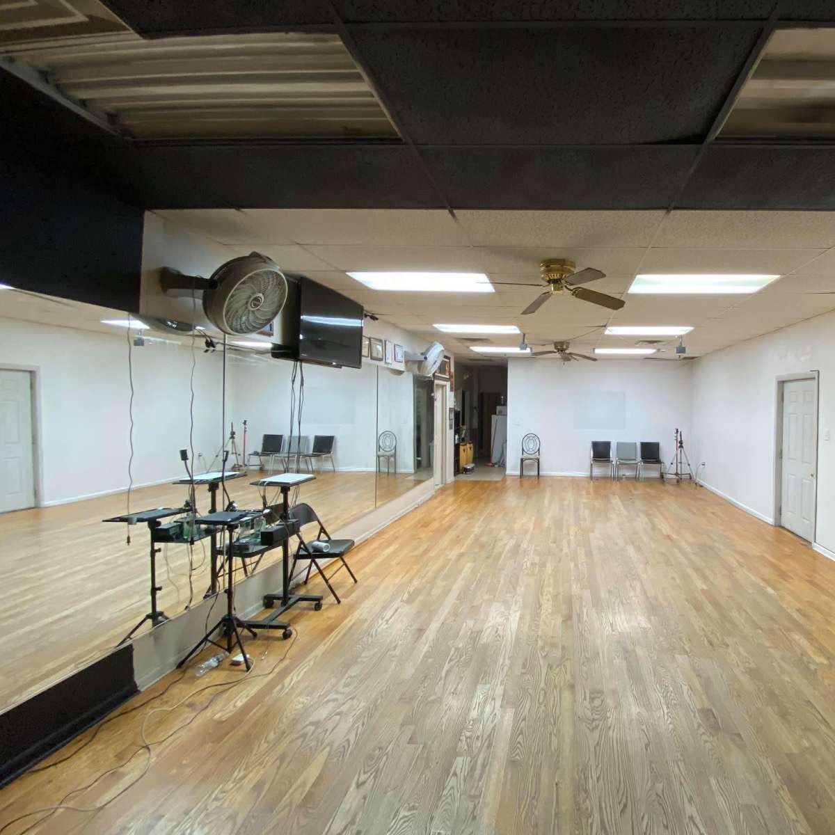 An empty room with wooden flooring, a ceiling fan, mirrors on one wall, and several chairs against the opposite wall.