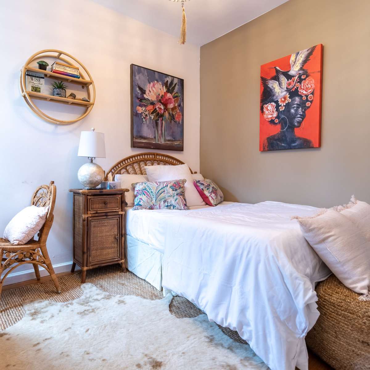 A small, decorated bedroom featuring a single bed with white bedding, a round wall shelf with plants, and a couple of artworks, alongside a wicker chair and a wooden bedside table.