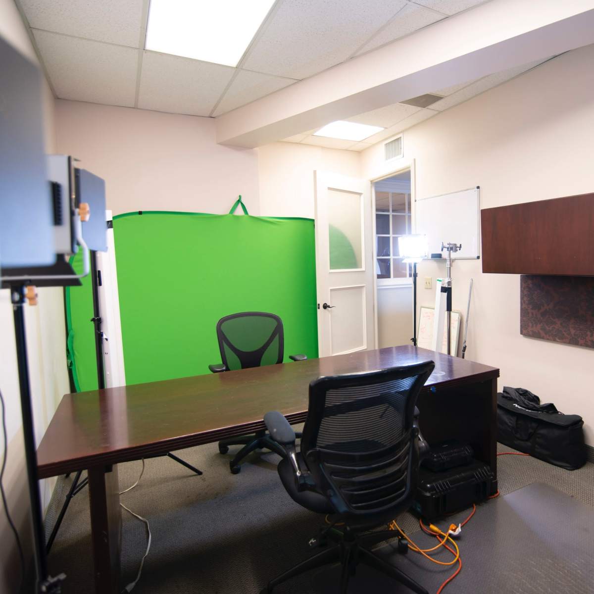 A sparsely furnished office featuring a wooden desk, a black office chair, and a green screen setup against the wall.