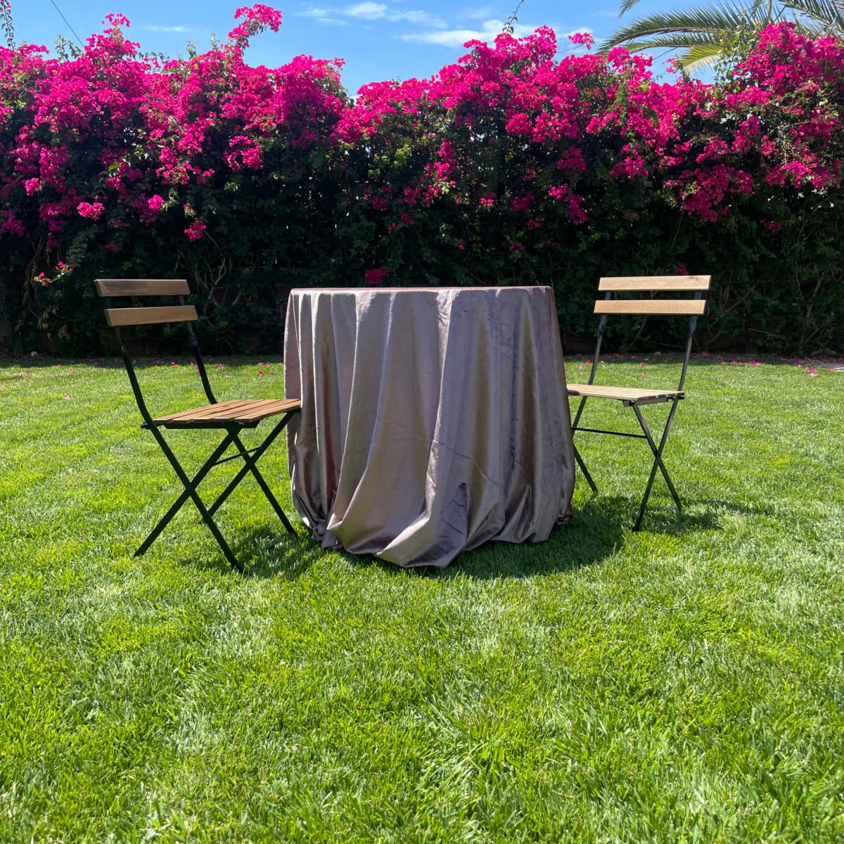 A round table with a tablecloth is set between two chairs on a grassy area, surrounded by blooming pink flowers and palm trees.