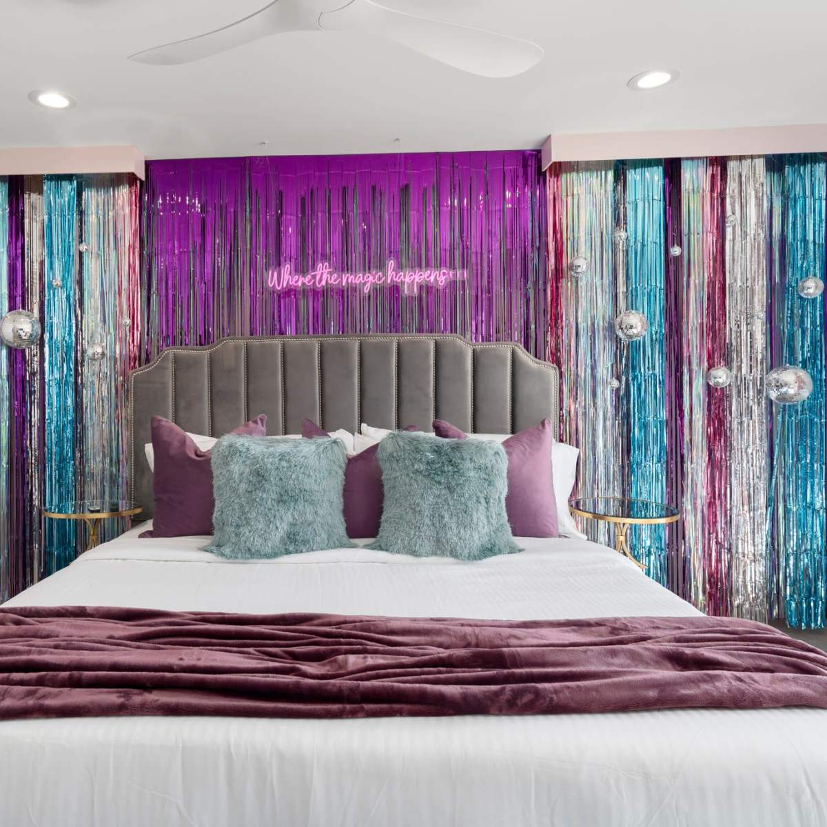 A modern bedroom with a bed featuring a gray headboard, adorned with purple and teal decorative elements, and a backdrop of shiny streamers and a neon sign.