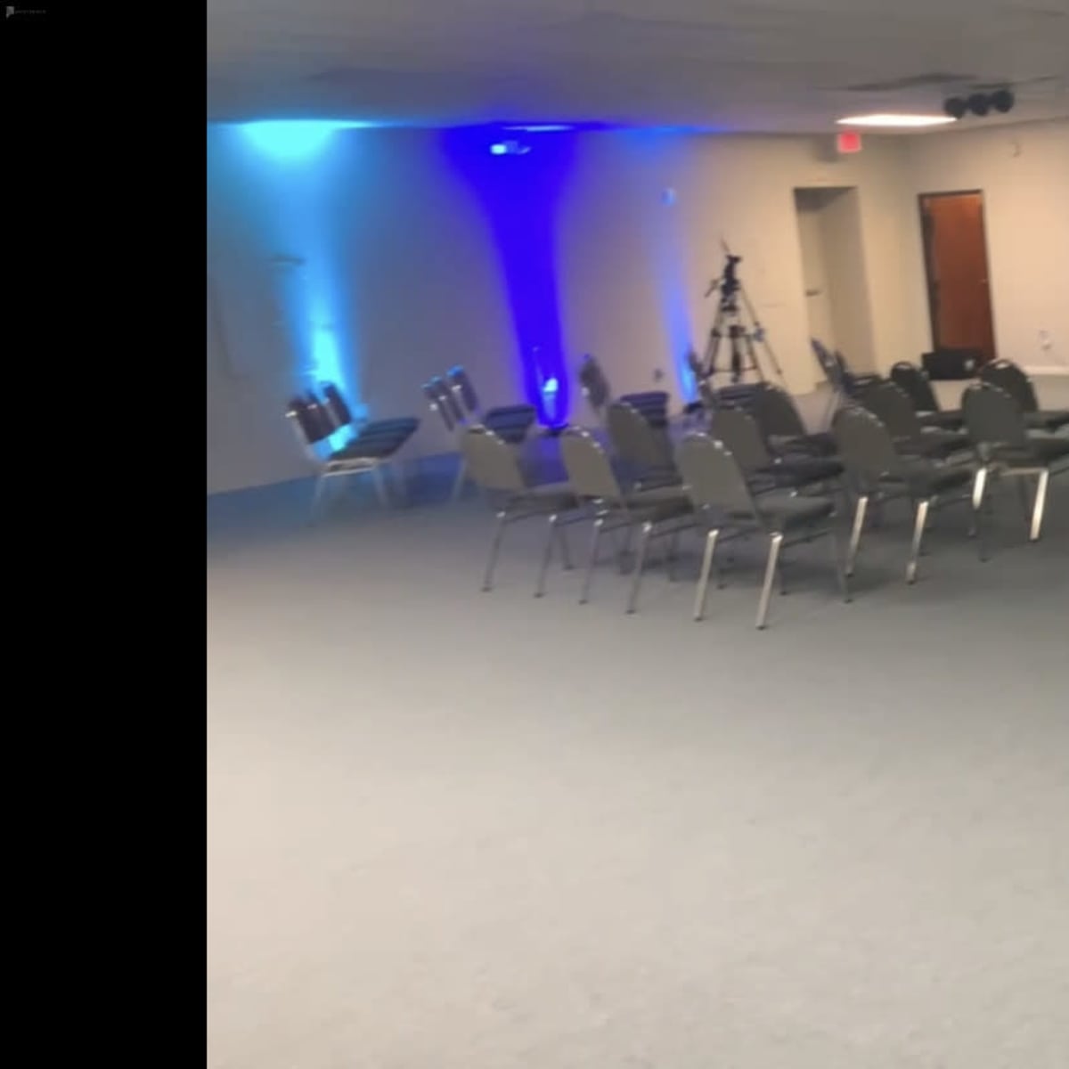 A vacant room set up for an event, featuring rows of metal chairs and a stage with lighting and sound equipment at the front.
