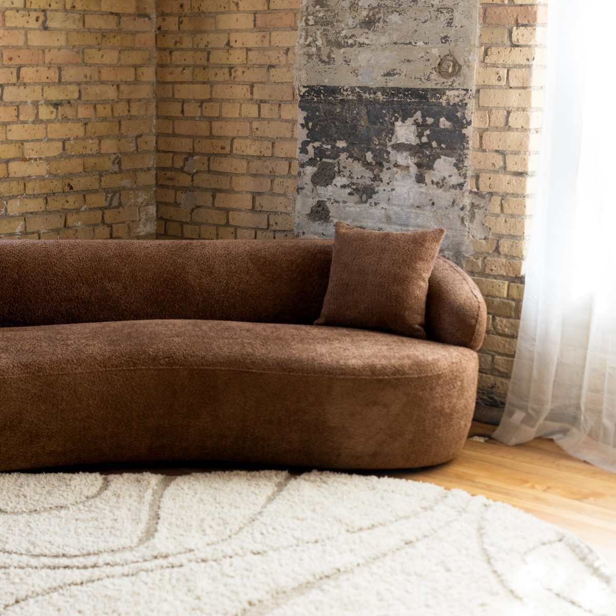 A brown, curved sofa with two matching cushions is positioned in a room with exposed brick walls and large windows draped with sheer curtains.