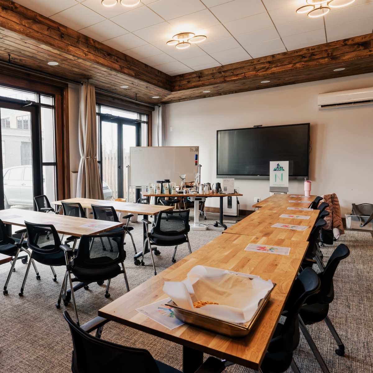 A spacious meeting room features several wooden tables arranged in a layout, with chairs positioned around them and a large screen mounted on the wall.