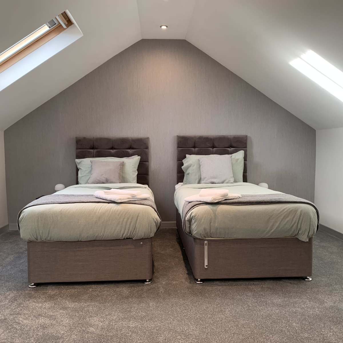 A well-lit attic bedroom with two single beds placed side by side, each featuring a gray upholstered headboard and light bedding.