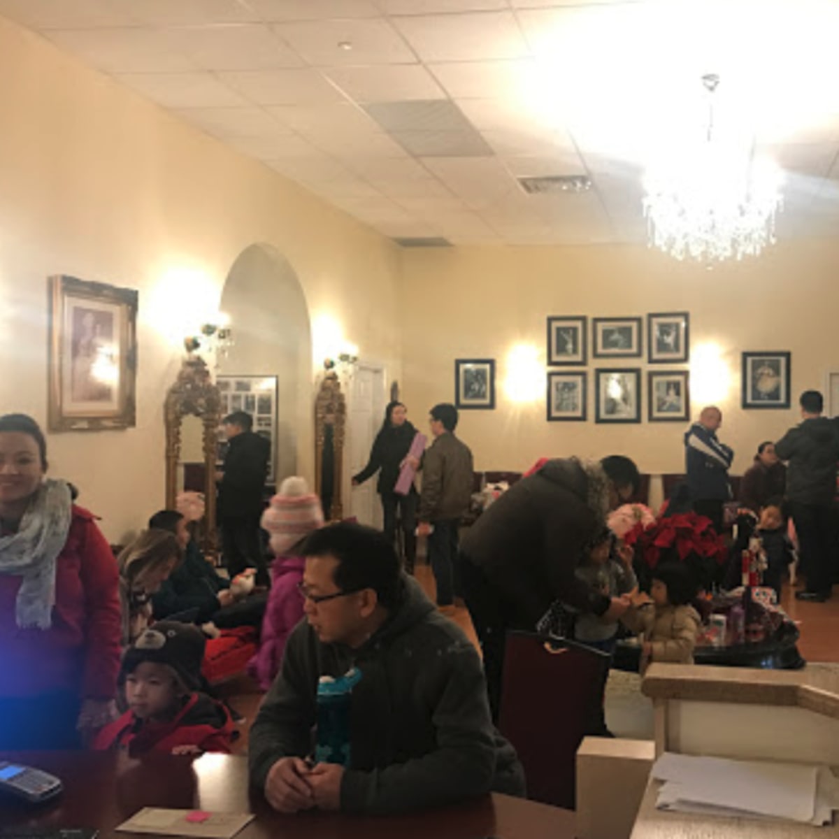 A group of people, including children, are gathered in a spacious room adorned with framed photographs and a chandelier, while some are seated and others are standing.