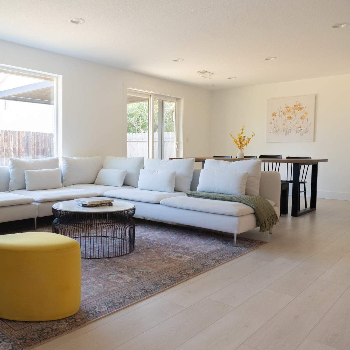 A light-filled living room features a sectional sofa with pillows, two yellow ottomans, a round coffee table, and a dining area in the background.