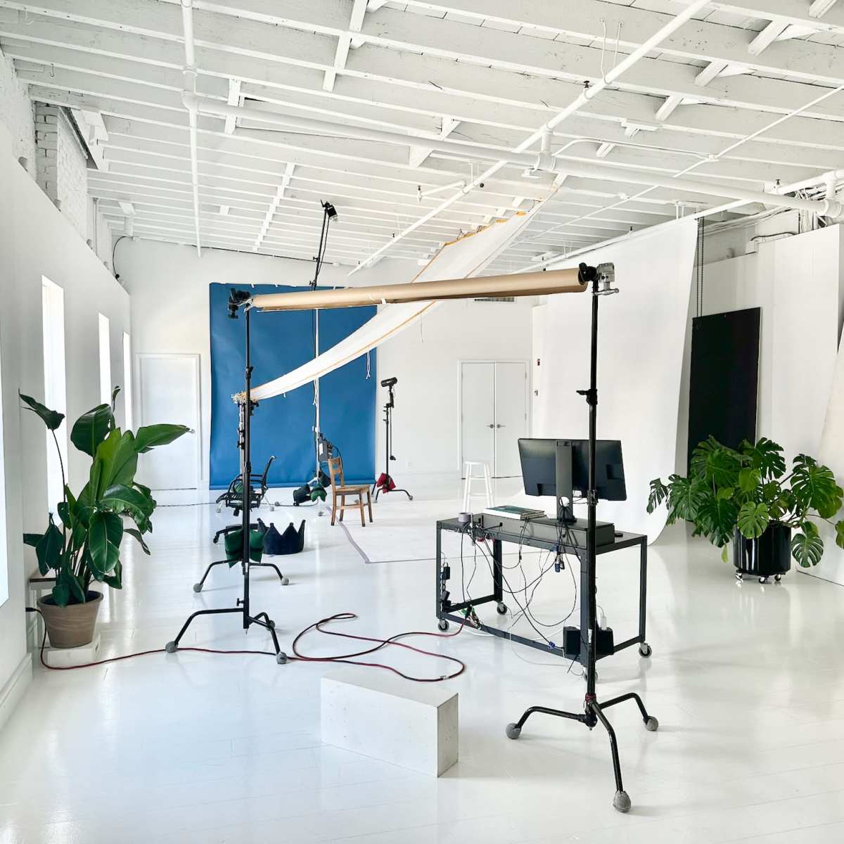 A bright studio space with white walls, a long table, potted plants, and equipment for photography, including backdrop supports and a computer setup.
