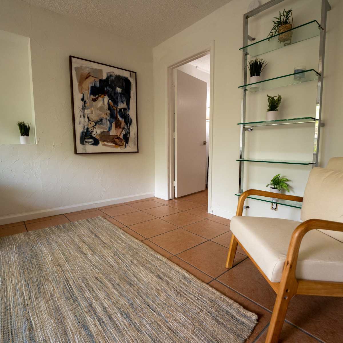 A minimalistic room features a beige armchair, a textured area rug, and a glass shelf with plants against a white wall.
