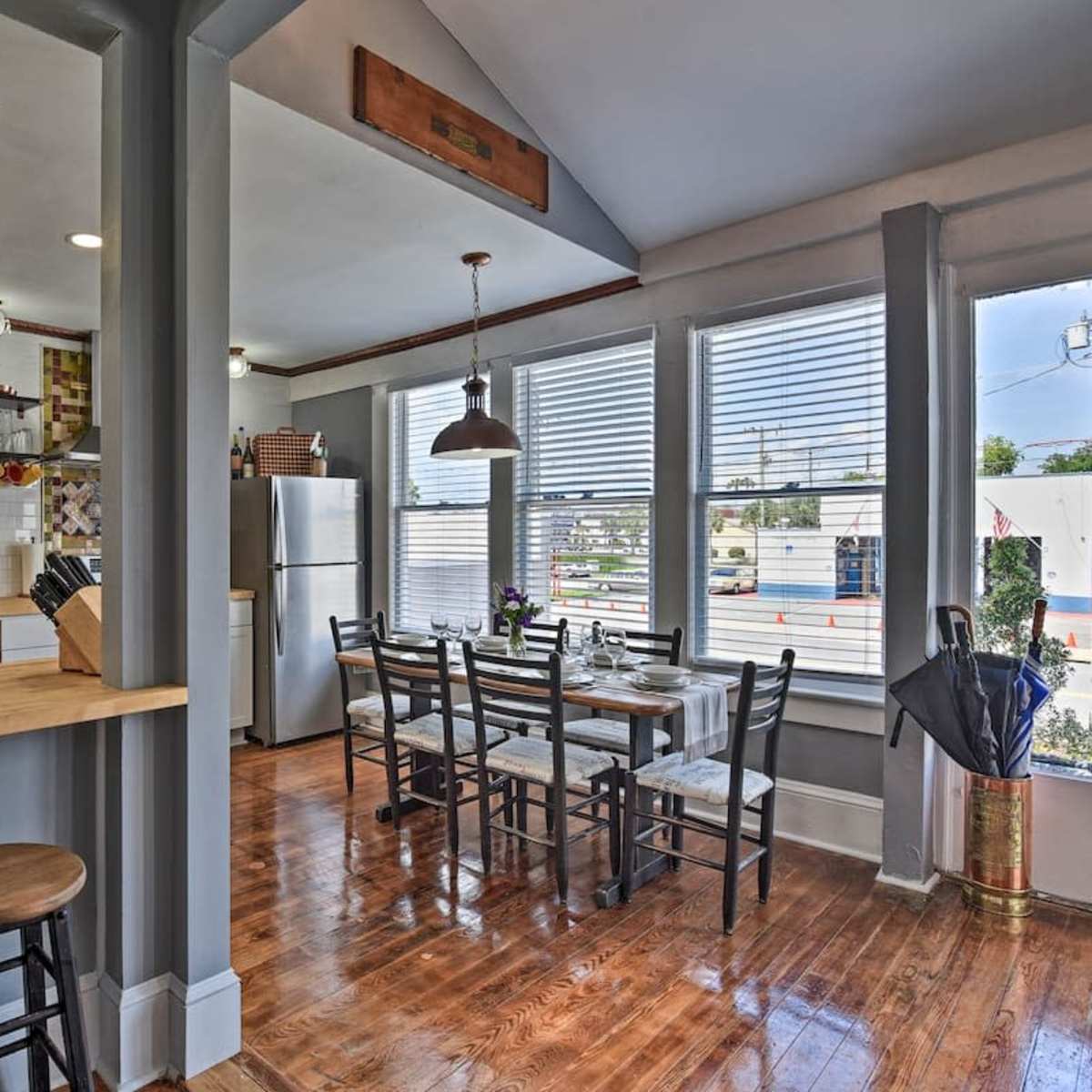 A kitchen and dining area feature wooden flooring, a bar counter with two stools, and a dining table set for four beneath large windows.