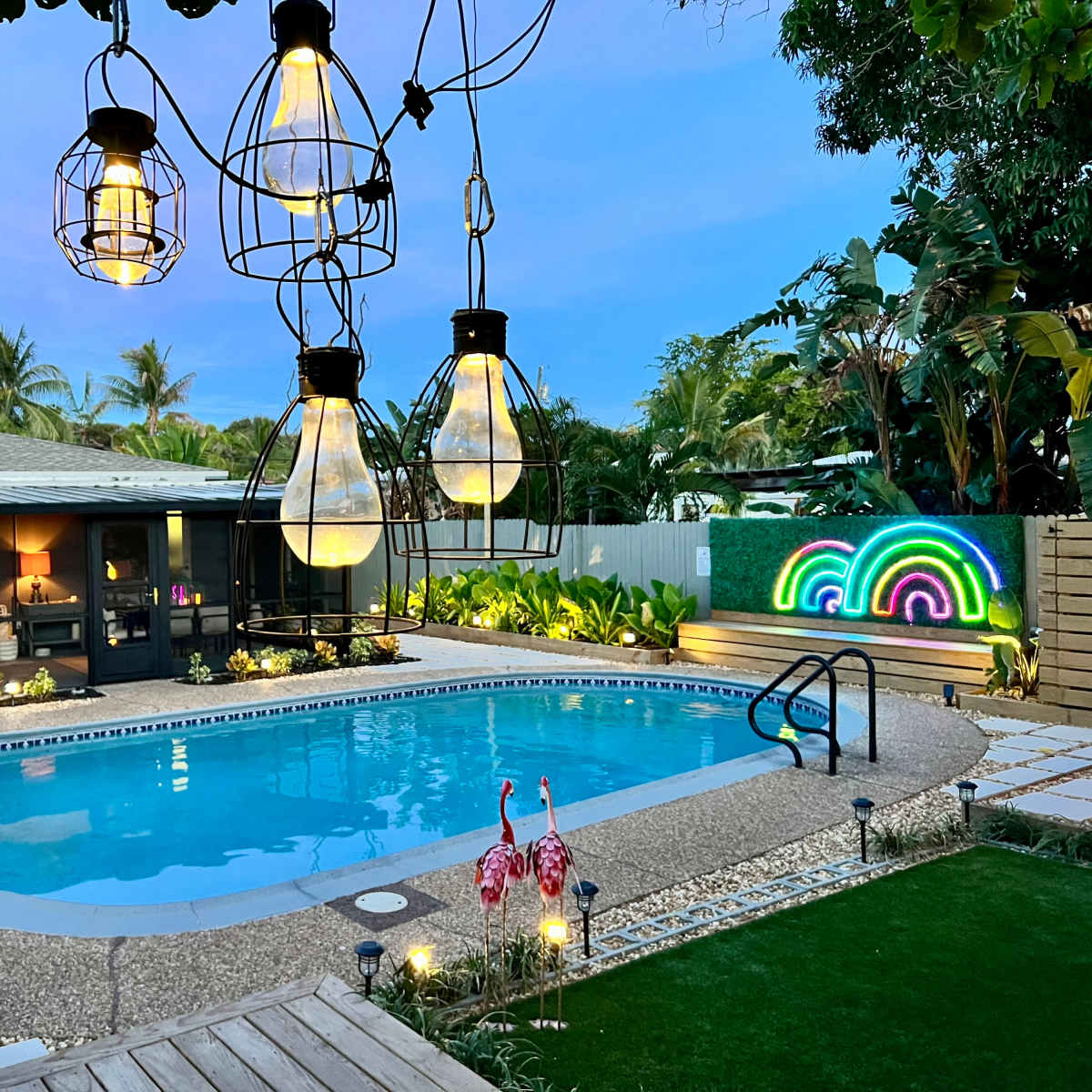 A backyard with a swimming pool surrounded by tropical plants, decorative lights hanging above, and a colorful light display on the wall.