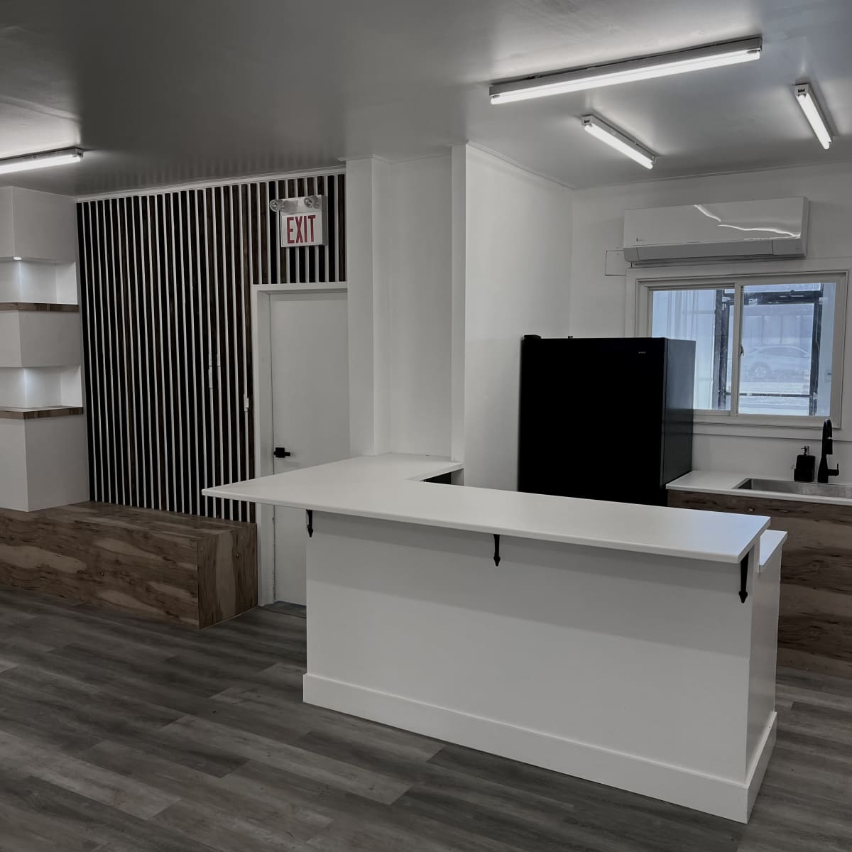 A modern interior space with a white service counter, wooden accents, and a kitchen area featuring a refrigerator and window.