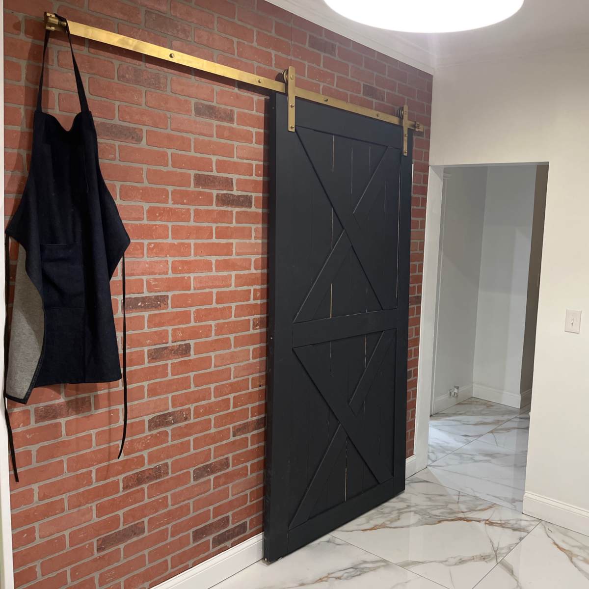 A kitchen area featuring a black sliding barn door on a brick wall, with an apron hanging beside it and white marble flooring.