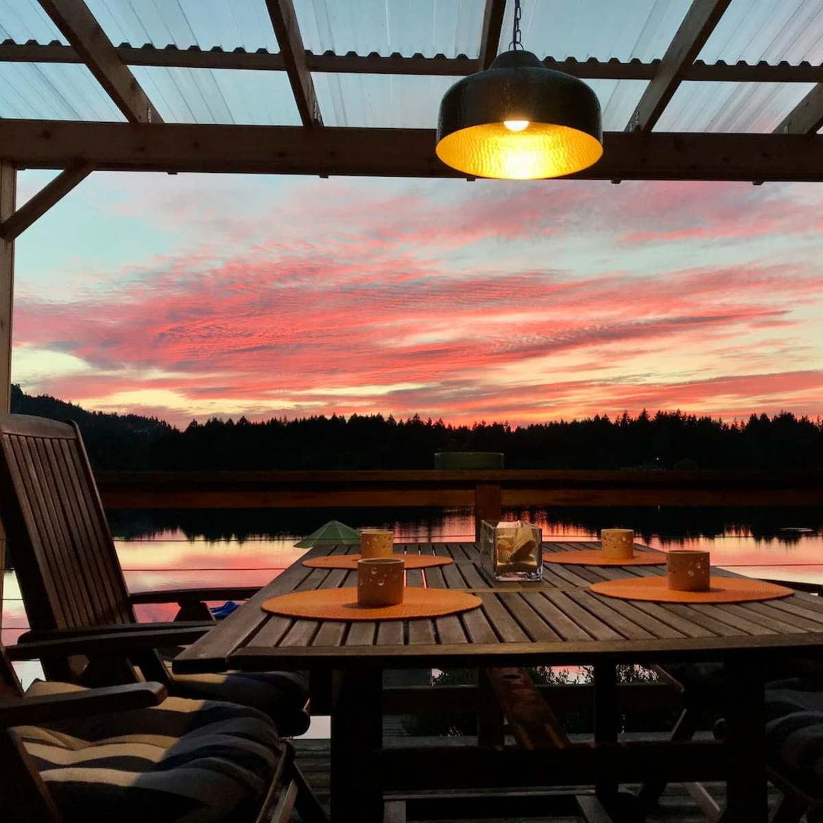 A wooden table and chairs are set under a covered patio, overlooking a calm lake at sunset, with vibrant colors in the sky reflected on the water.