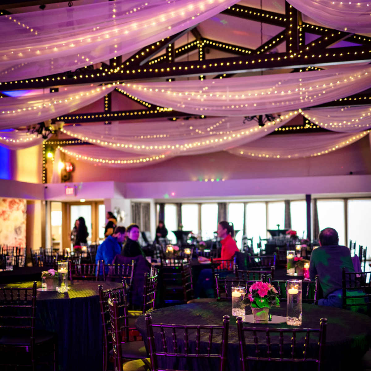 The interior space features round tables with black tablecloths, surrounded by chairs, adorned with soft lighting and draped fabric overhead.
