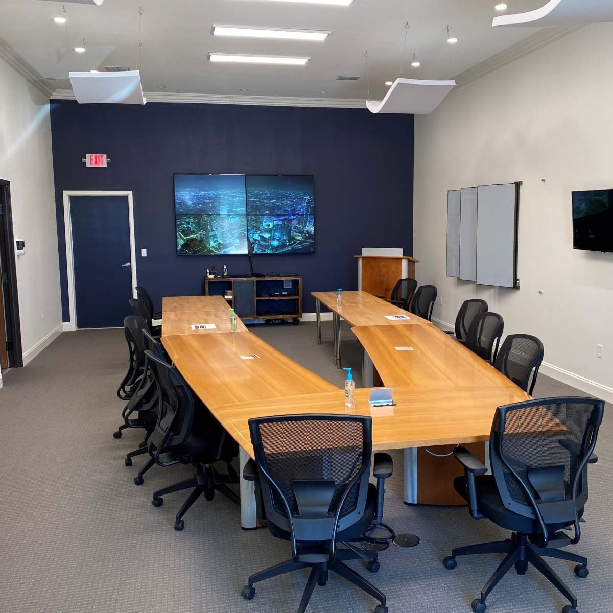 A modern conference room with a long wooden table, black office chairs, and multiple screens mounted on the walls.