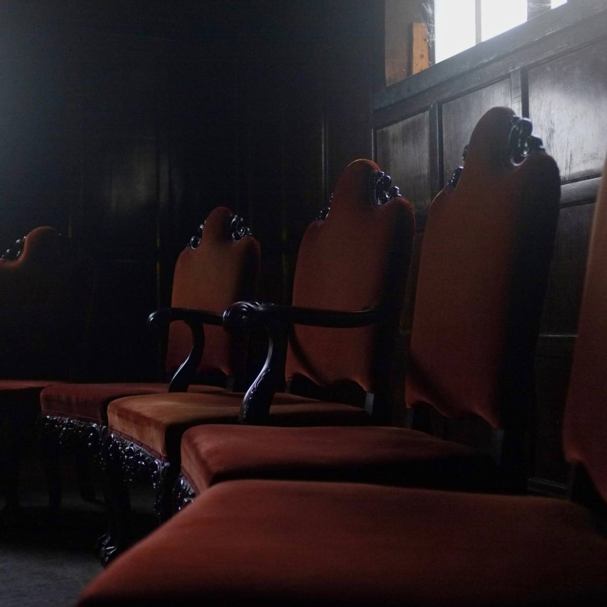 A row of ornate, upholstered chairs is positioned in a dimly lit room with a small window.