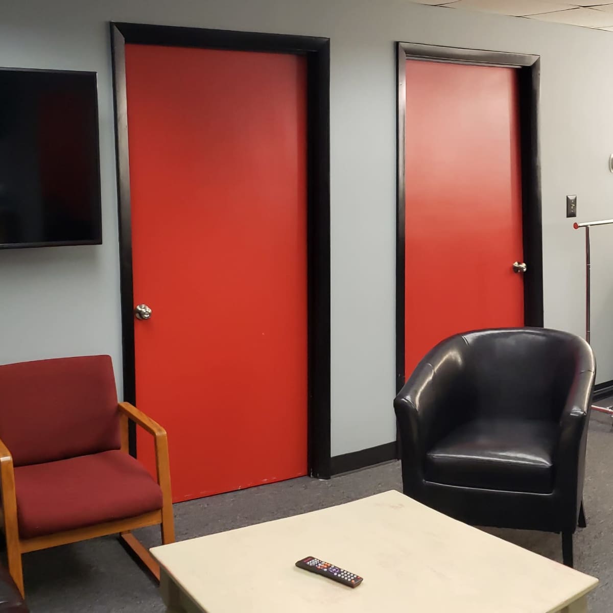 A lounge area with two red doors, a black sofa, several black chairs, a bar-style counter with high stools, and a television mounted on the wall.