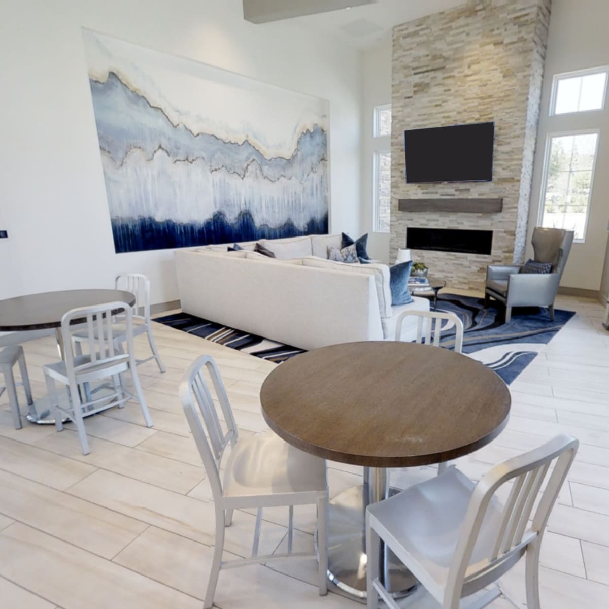 A modern lounge area with a large painting, a light-colored sofa, and two small tables with chairs, featuring large windows that allow natural light in.