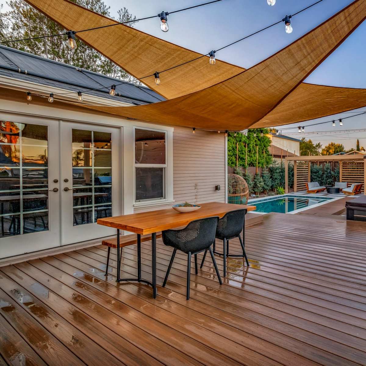 A wooden deck features a dining table with chairs, a pool in the background, and a shade structure overhead, accompanied by string lights.