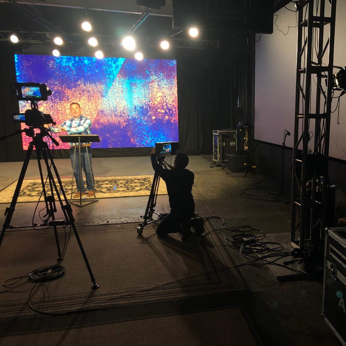 A speaker stands at a podium in front of colorful screens, while cameras are set up to capture the presentation.