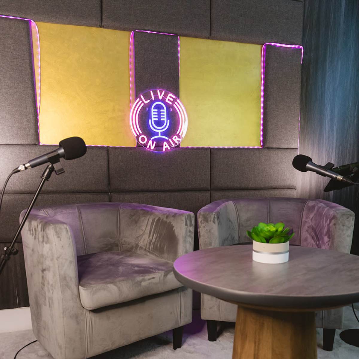 A small podcast studio setup with two grey chairs, a wooden table, and a neon sign that reads "LIVE ON AIR" against a textured wall.
