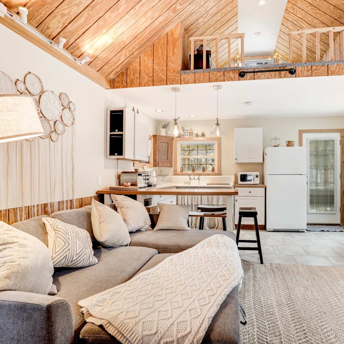 A cozy living space featuring a gray sofa, a kitchenette with a white refrigerator, and wooden accents on the walls and ceiling.