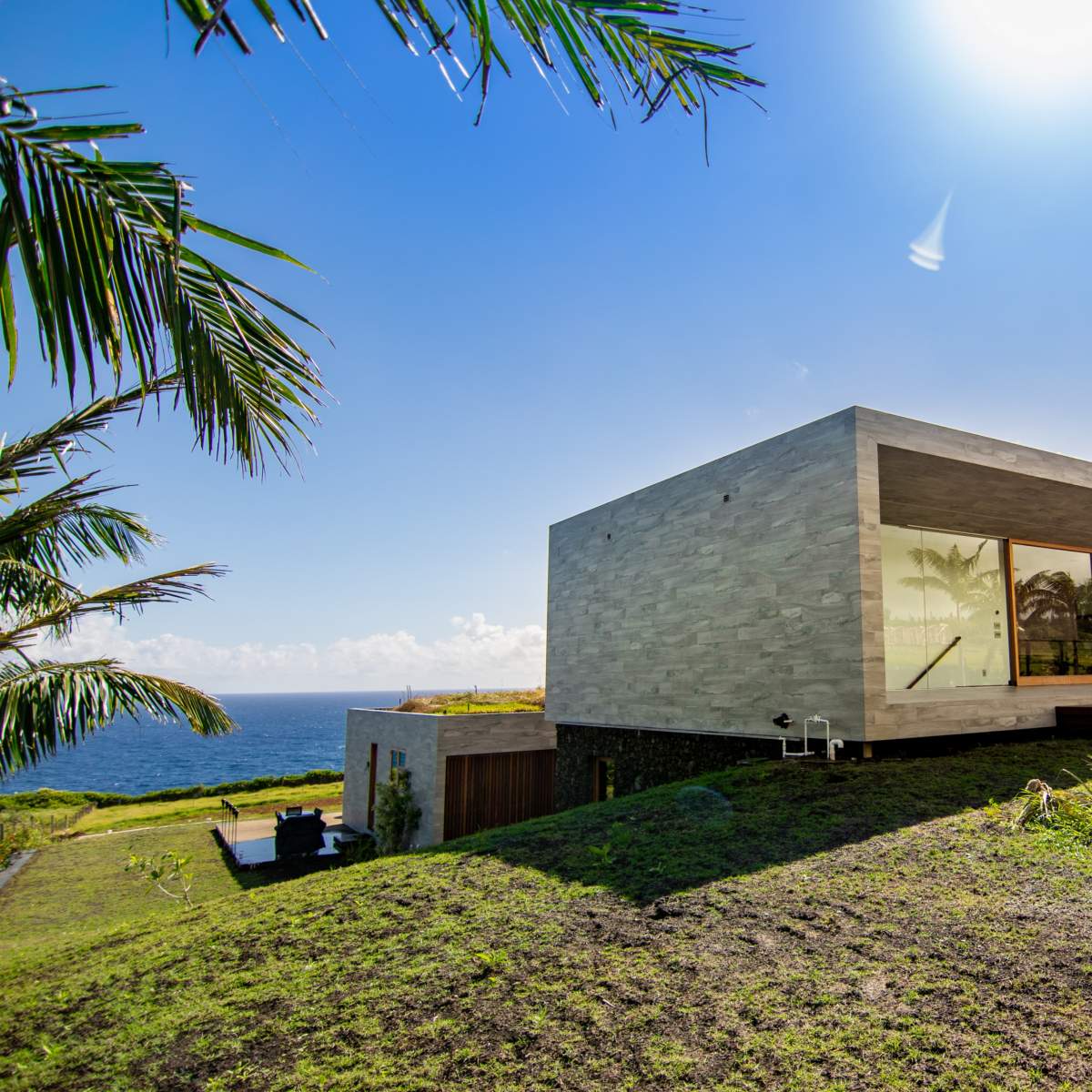 A modern concrete house with large windows sits on a grassy hillside overlooking the ocean.