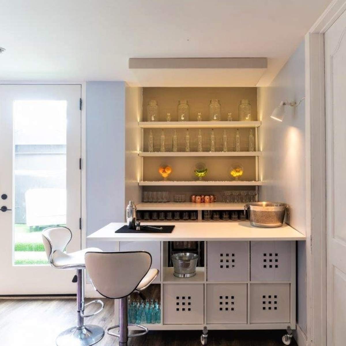 A modern kitchenette with open shelving displaying glass containers and a small bar area equipped with a sink and seating, beside a set of double doors leading to an outdoor space.