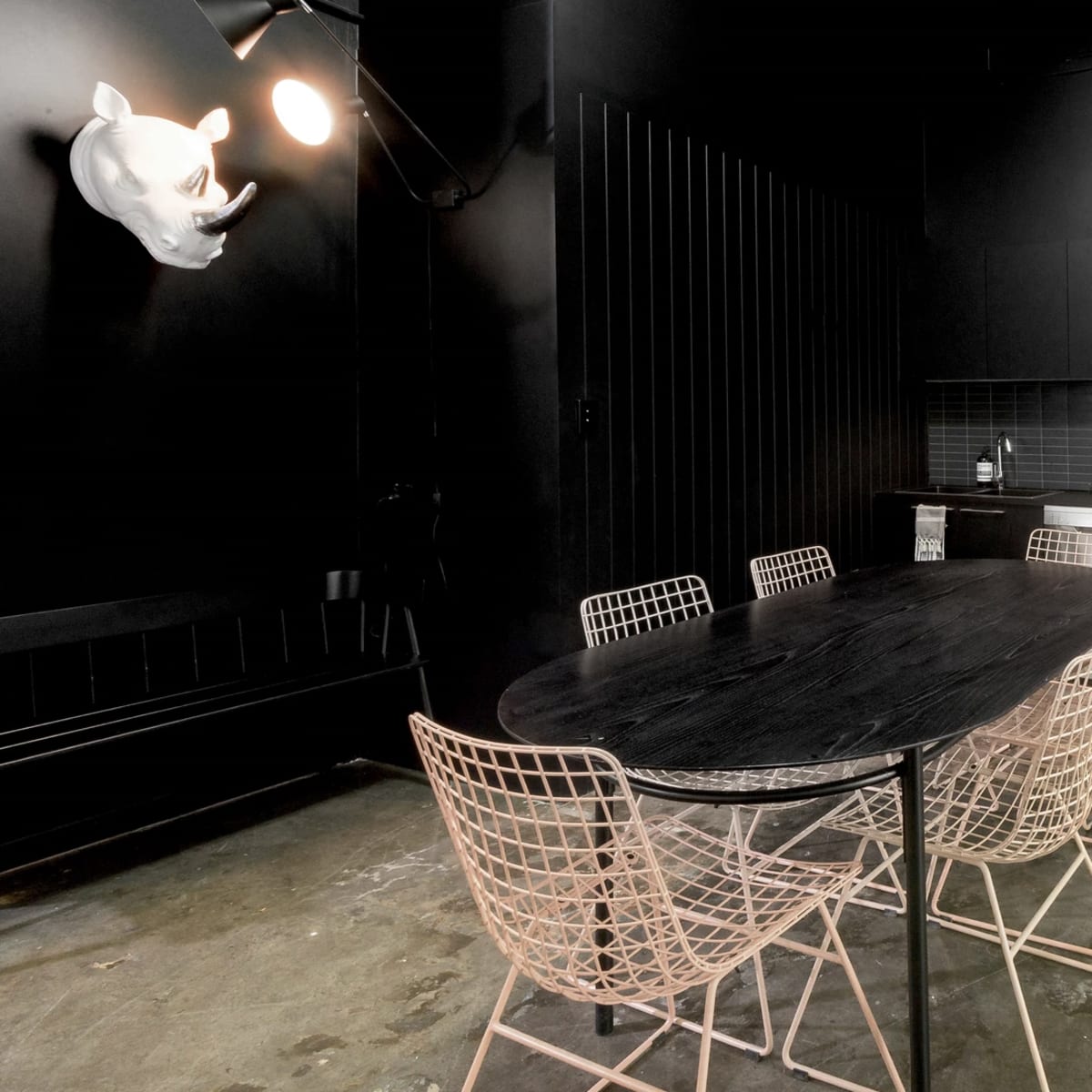 A modern kitchen space with a large black table surrounded by wire chairs, featuring dark walls and a sleek counter with appliances and a mini fridge.