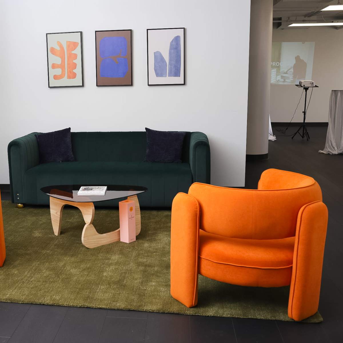 A seating area features a green couch, two orange armchairs, a wooden coffee table, and three framed artworks on the wall, with a potted plant in the corner.