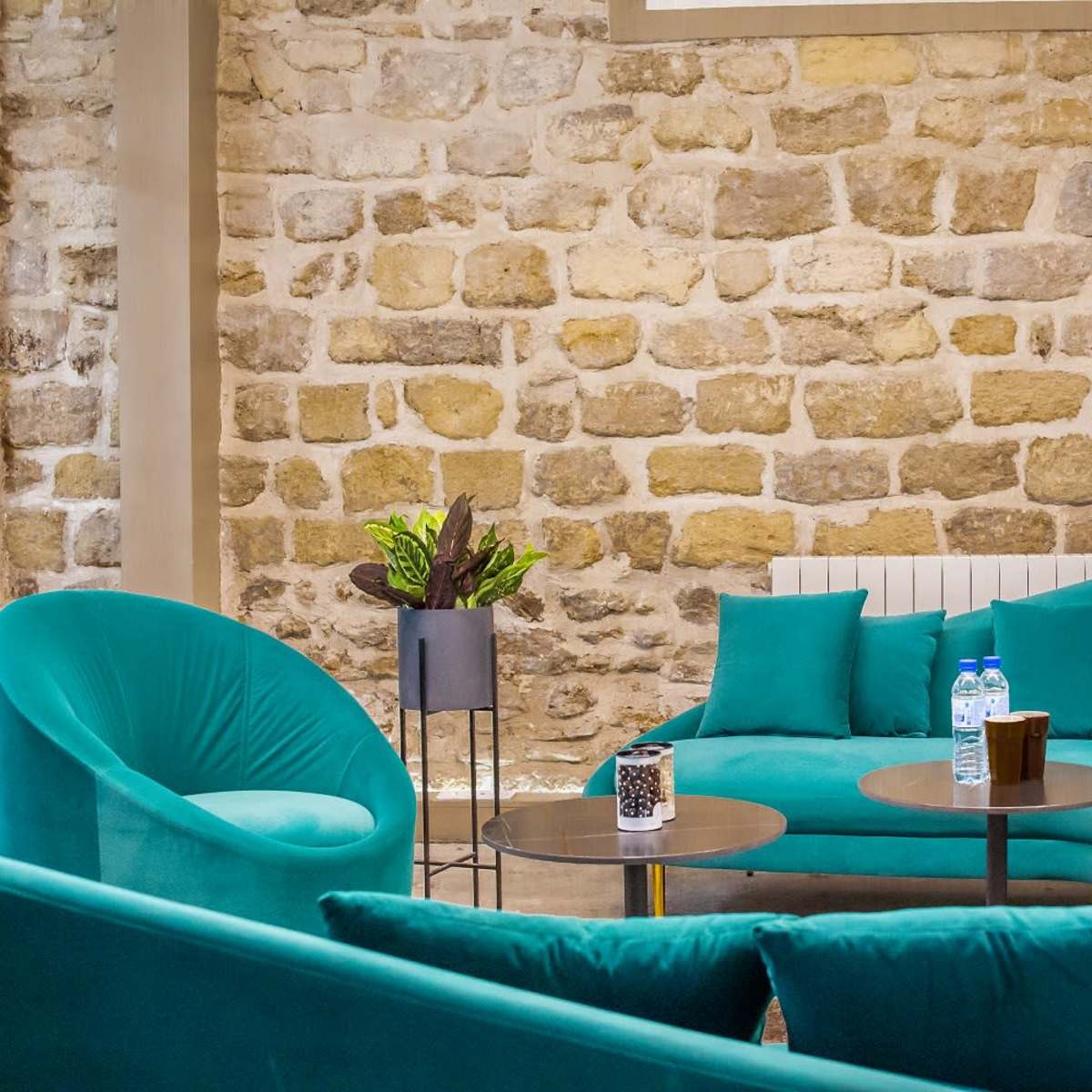 A cozy seating area with teal, curved sofas, small black tables, and potted plants against a stone wall backdrop.